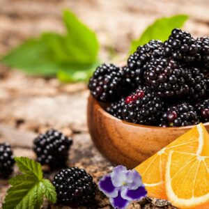 A wooden bowl filled with fresh blackberries, accompanied by a sliced orange and green mint leaves, set on a rustic wooden surface, infused with Dark Cassis Fragrance Oil for use in candle making, soap making, perfumes, diffusers and more.