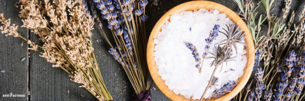 A panoramic image of a wooden bowl filled with white bath salts and lavender stems, set against dried lavender bouquets on a rustic gray wooden background.