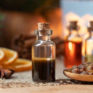 A close-up of a small glass bottle with Oriental Myrrh & Musk fragrance oil for use in candle making, soap making, perfumes, diffusers and more., sealed with a cork, on a rustic table with blurred bottles, orange slices, and raisins in the background.