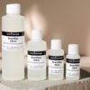 Image showing four bottles of various sizes, all labeled "ScentStay" by zenAroma. The bottles, ranging from 30ml to 250ml, stand on a textured slab against a neutral background. Rose petals are scattered to the side, adding a touch of color to the minimalist setting, emphasizing ScentStay's role as a fragrance fixative.