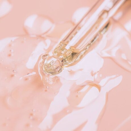 Close-up of a dropper releasing ScentStay onto a smooth, light pink surface. The liquid appears slightly viscous and forms small bubbles and ripples around the dropper tip. The scene has a soft, pastel aesthetic.